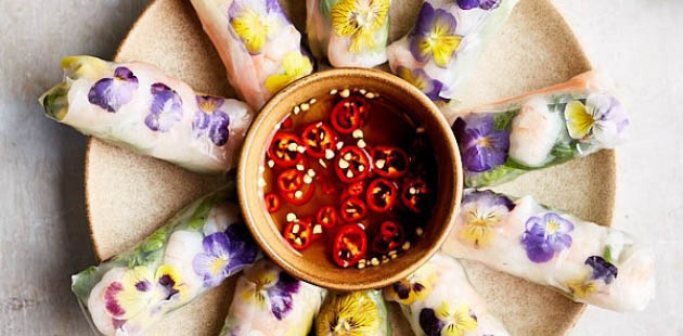 A plate of spring rolls with purple and yellow petals are pressed into the rice paper. In the centre is a bowl full of chilli oil to dip the spring rolls in.
